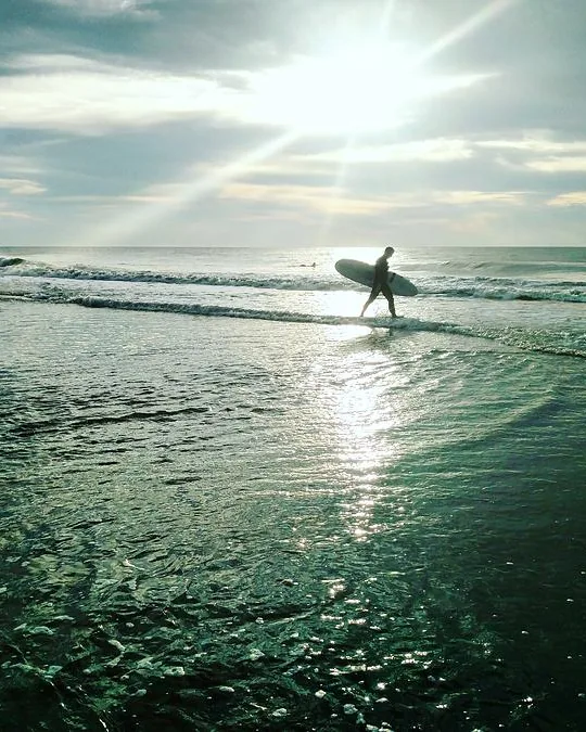 A surfer walks out of the sea into low sun