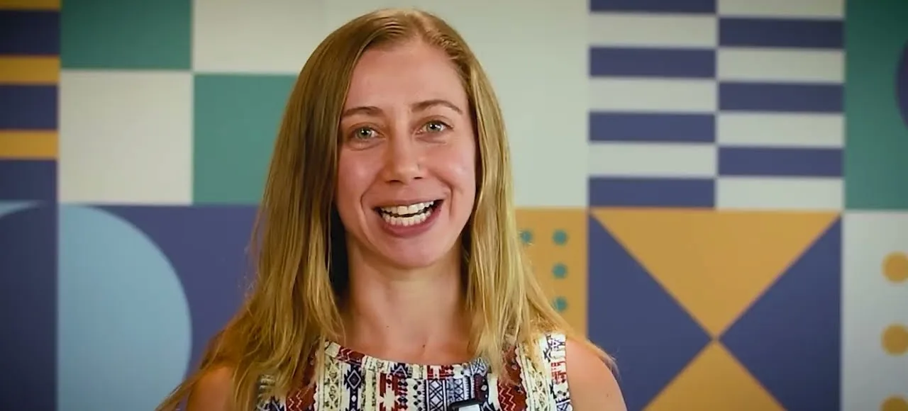 A presenter smiles at the camera in front of a bright geometric wall
